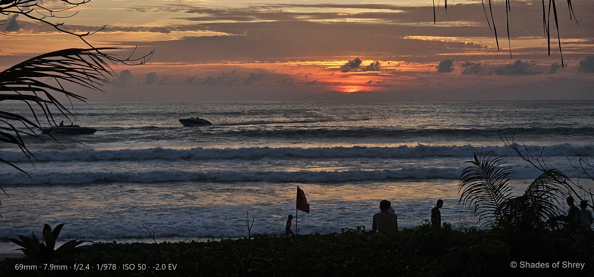 Golden sunset over ocean waves with boats and palm silhouettes, people watching from shore