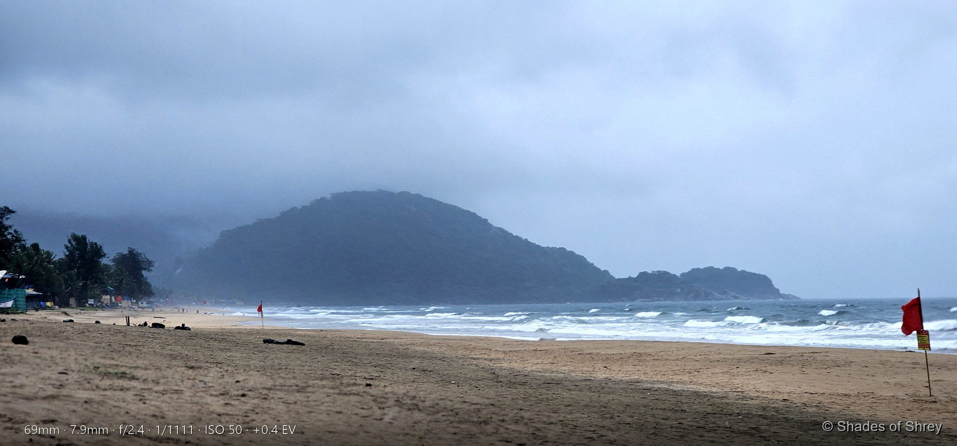 Misty monsoon beach with green headland emerging from clouds, red warning flags, Goa
